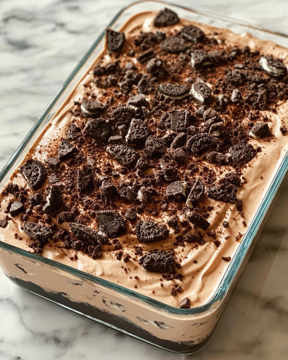 A glass rectangular dish filled with a creamy dessert that has two visible layers, the bottom layer being a smooth, light brown base and the top layer covered with a thick, lighter brown mousse or cream. The top is generously sprinkled with large and small chunks of dark brown chocolate cookies, creating a rough texture and contrast. The dish is placed on a white marbled surface, enhancing the rich colors of the dessert. photo taken with an iphone --ar 4:5 --v 7