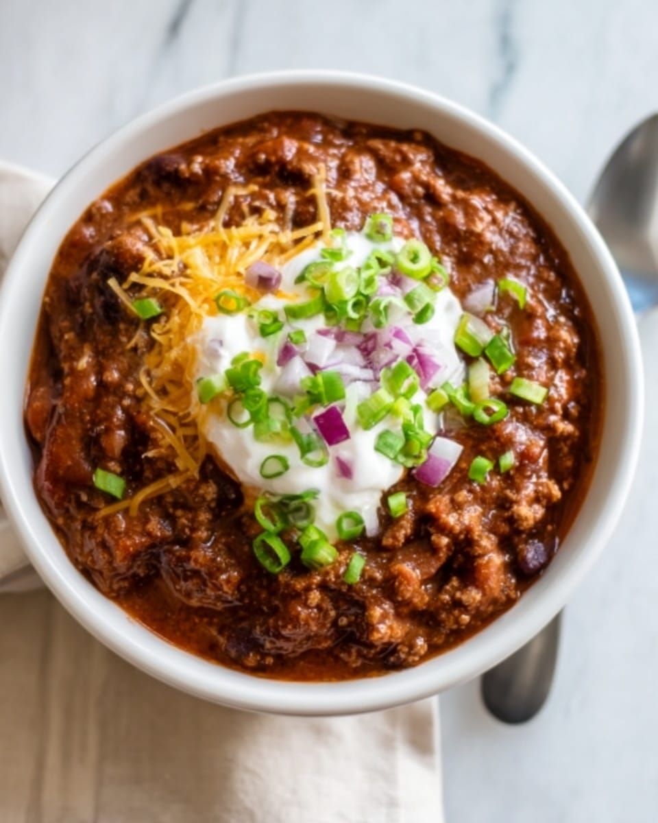 A white bowl filled with a thick layer of chili, showing a rich brown color with visible ground meat and beans. On top, there is a dollop of white sour cream, sprinkled with finely chopped green onions and small pieces of red onion. A small amount of shredded yellow cheese is also visible peeking through the chili near the edges. The bowl is placed on a white marbled surface with a white spoon next to it. Photo taken with an iphone --ar 4:5 --v 7