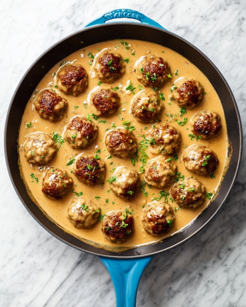 A black skillet with a blue handle is filled with two layers: the bottom layer is a smooth, creamy light brown sauce, and on top are about twenty round meatballs browned on the outside and mostly covered in the same sauce. Small green herb pieces are sprinkled on top of the meatballs and sauce, adding a fresh contrast. The skillet sits on a white marbled surface. photo taken with an iphone --ar 4:5 --v 7
