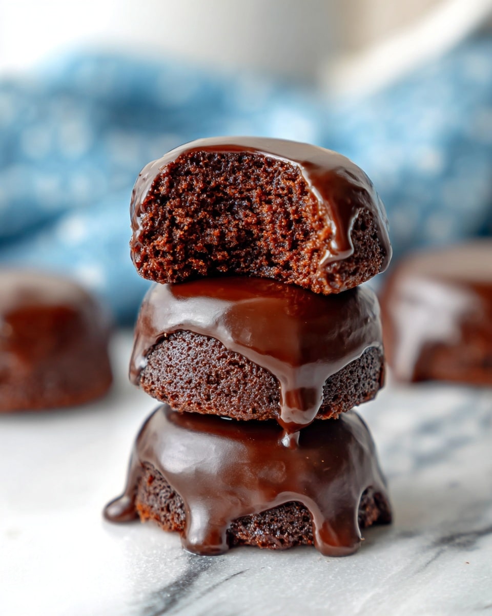 A stack of three small, round chocolate cakes covered in smooth, shiny dark chocolate glaze is shown, with the top cake bitten to reveal its moist, dense, dark brown crumb texture inside; the glaze gently drips slightly down the sides of each cake, giving a rich, glossy finish, all set on a white marbled surface with a blurred background featuring hints of blue and white. Photo taken with an iphone --ar 4:5 --v 7