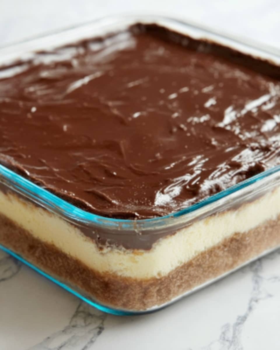 The image shows a glass square dish filled with a layered dessert topped with a smooth, shiny layer of chocolate spread evenly across the top. The dessert appears to have multiple layers visible through the glass sides: a bottom crust layer in a light brown color, a thicker creamy beige layer in the middle, and the glossy dark brown chocolate layer on top. The dish is placed on a white marbled surface, and the edges of the glass dish are clear with a slight blue tint. Photo taken with an iphone --ar 4:5 --v 7