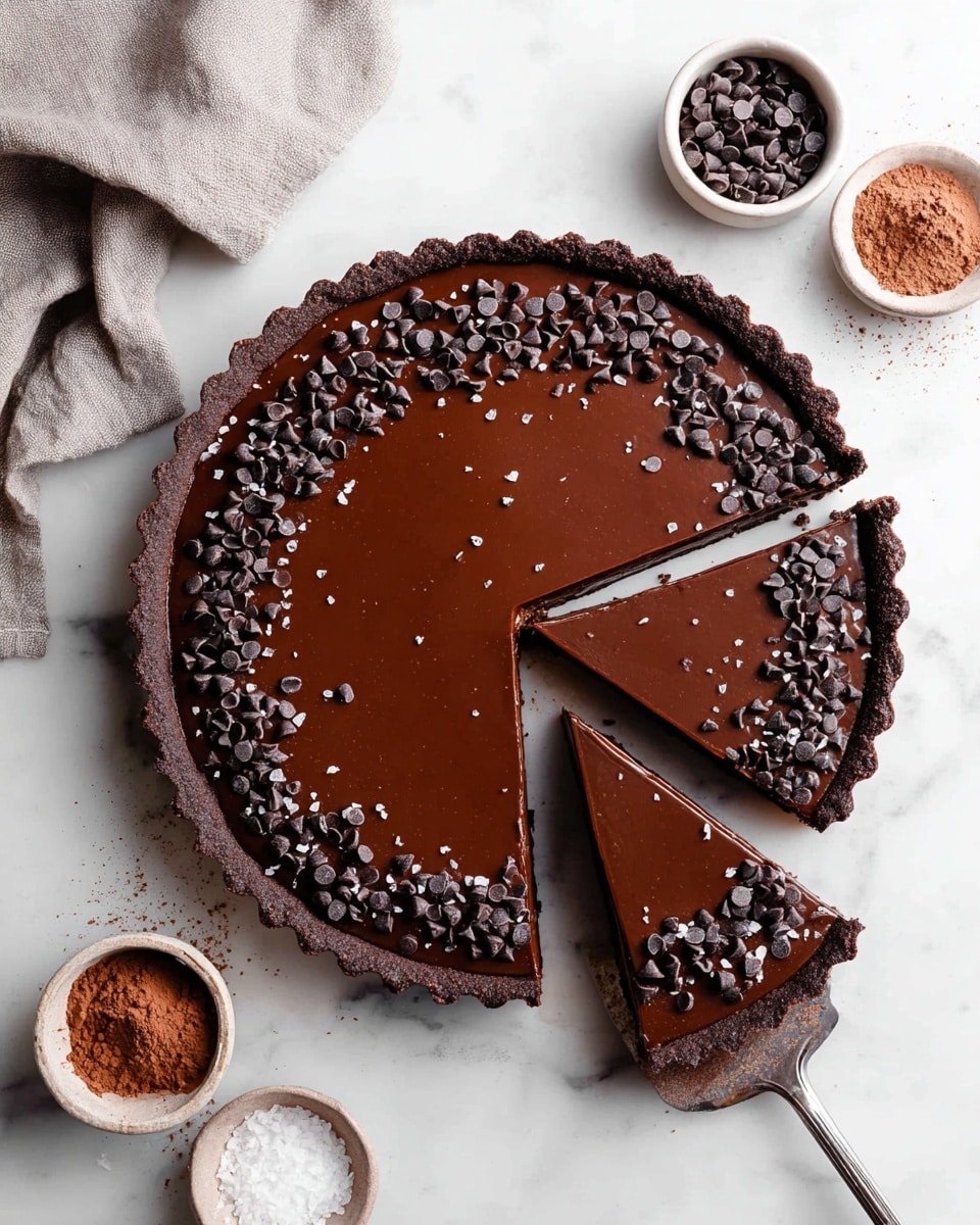 A round chocolate tart with a dark brown crust forms the base, filled with a smooth, glossy chocolate layer on top. Around the edge of the chocolate filling, there is a ring of small dark chocolate chips sprinkled with coarse sea salt flakes. Three slices have been cut and slightly pulled out on the right side, with a silver pie server visible under one slice. The tart sits on a white marbled surface, surrounded by small white bowls containing cocoa powder, sea salt, and more chocolate chips. A light gray cloth is draped in the top left corner. photo taken with an iphone --ar 4:5 --v 7