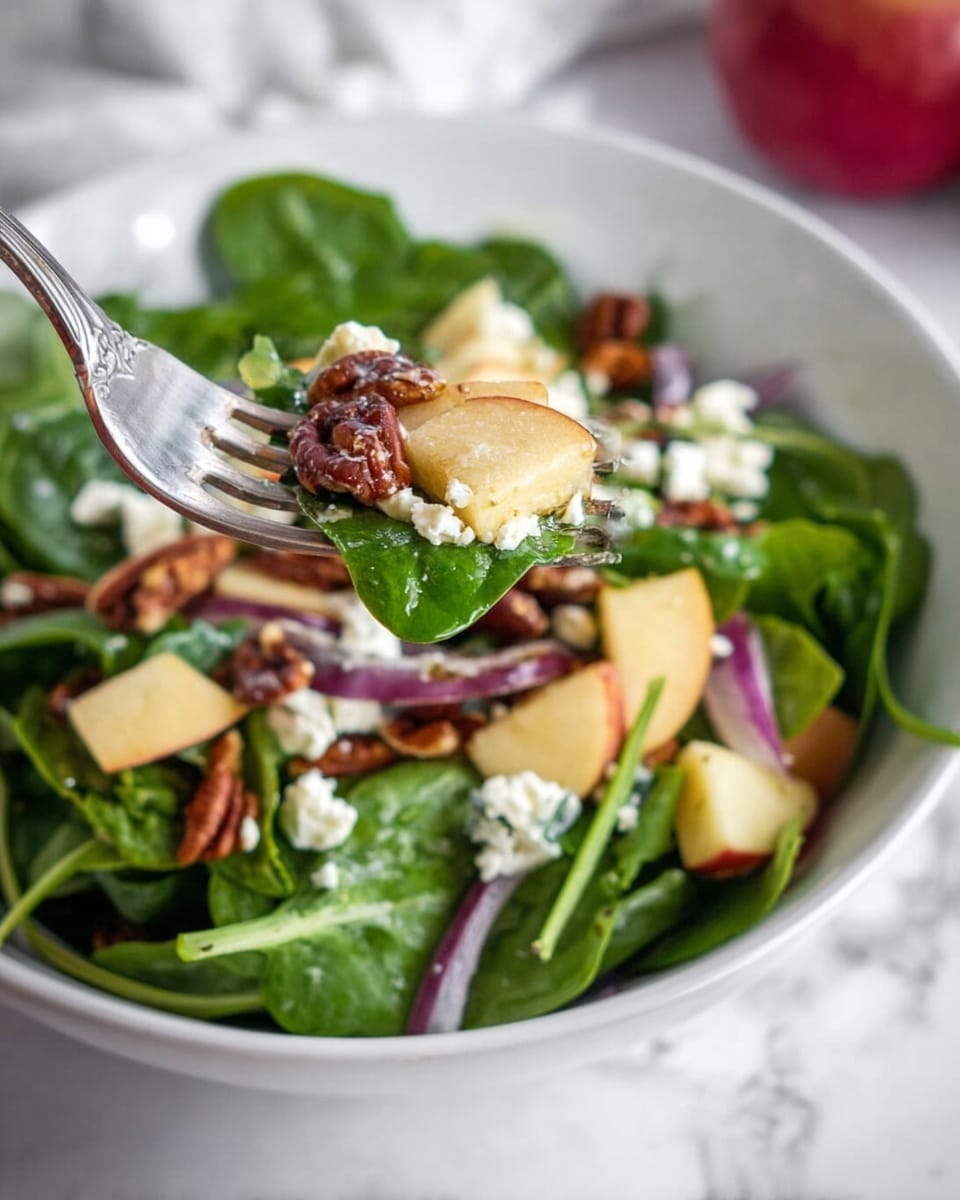 A white bowl contains a fresh salad with three main layers: bright green spinach and arugula leaves fill the base with a crisp and leafy texture, above them are chunks of light yellow apple pieces and purple-red onion slices adding color and crunch, and scattered on top are brown toasted pecans and white crumbly feta cheese providing texture and contrast. A silver fork lifts a forkful showing all these layers together against a white marbled surface in soft natural light. Photo taken with an iphone --ar 4:5 --v 7
