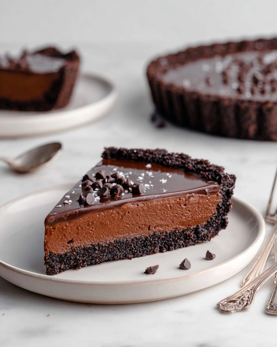 The image shows a slice of rich dark chocolate tart placed on a simple white plate against a white marbled background. The tart has three layers: a dark, crumbly chocolate crust on the bottom with a rough texture, a thick middle layer of creamy, smooth chocolate filling that is slightly lighter in color, and a glossy, shiny dark chocolate ganache layer on top. The ganache is topped with scattered dark chocolate chips and a few specks of flaky salt. In the background, there is another slice and the matching whole tart in a fluted tart pan, both with the same layers and texture. A pair of ornate silver utensils is partially visible on the right. photo taken with an iphone --ar 4:5 --v 7