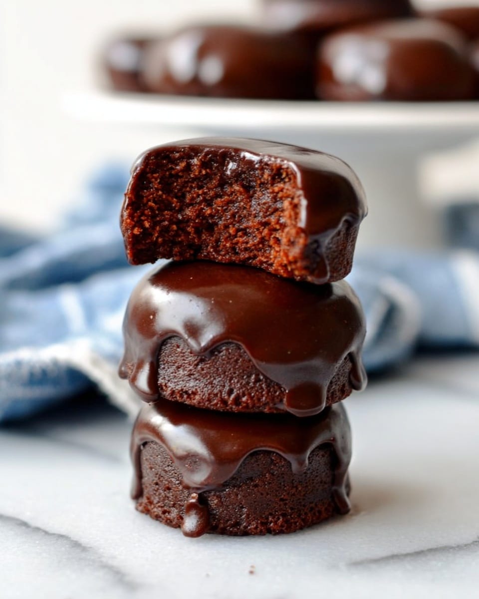 The image shows a stack of three small, round chocolate cakes covered in thick, glossy dark chocolate glaze. The bottom two cakes are whole, with the top cake partially bitten, revealing a moist, rich, and crumbly chocolate interior. The glaze drips slightly down the sides, adding a shiny texture to the dark brown layers. The stack is placed on a white marbled surface with a soft-focus white plate holding more cakes in the background, along with a blurred blue and white cloth. Photo taken with an iphone --ar 4:5 --v 7