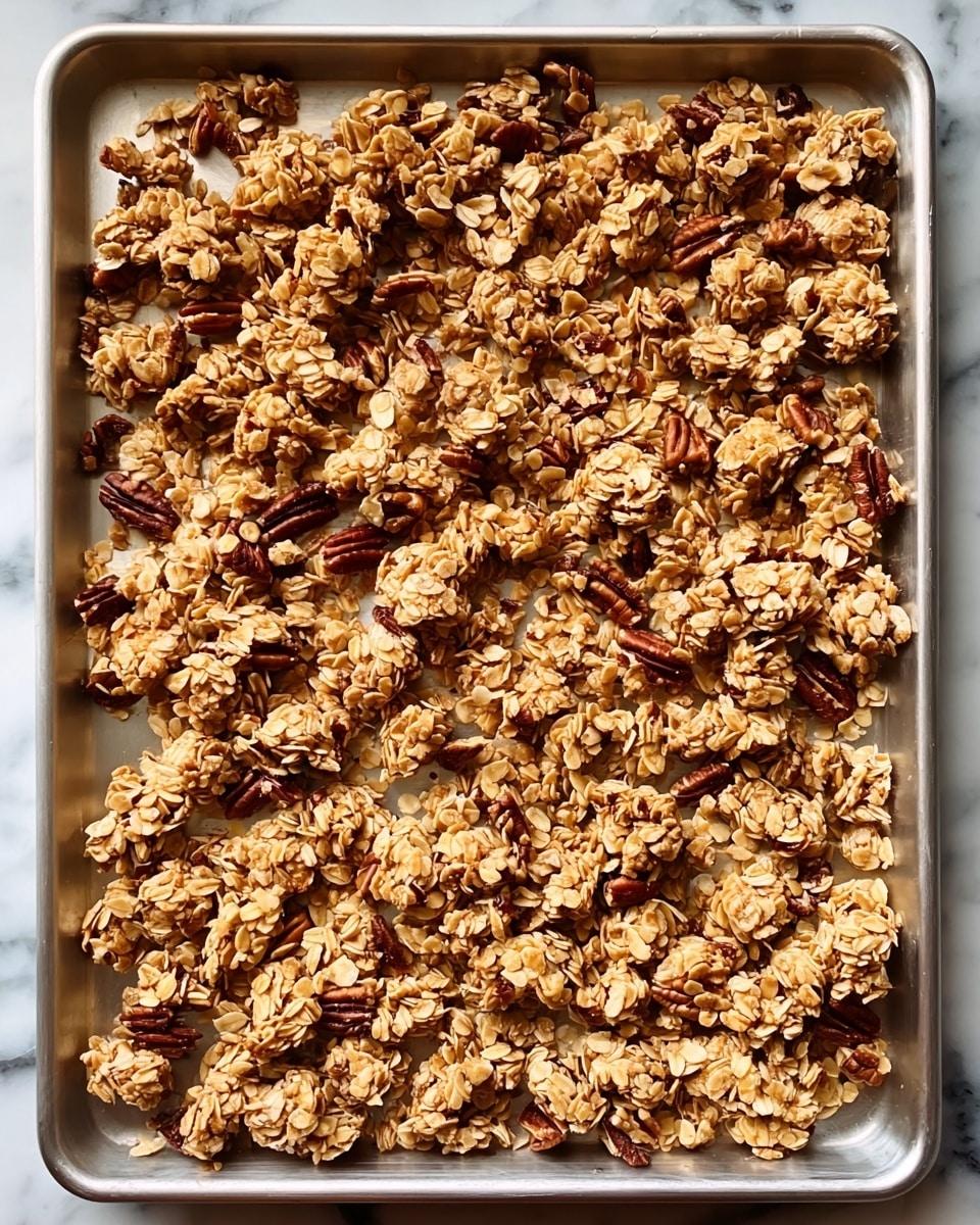 A tray filled with many small clusters of oat granola scattered evenly, showing a rough texture of oats mixed with nuts and seeds. The oats are light brown with some darker toasted patches, mixed with dark brown pecan pieces, giving a crunchy appearance. The tray is metal and the surface underneath it is white marbled texture. photo taken with an iphone --ar 4:5 --v 7