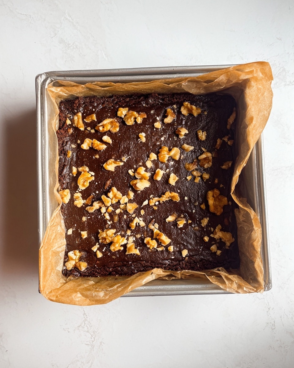 A square metal baking tray lined with crumpled parchment paper holds a thick, dark brown brownie layer that fills the tray nearly edge to edge, topped with scattered light brown chopped walnuts. The brownie surface looks moist and slightly glossy with a few small cracks. The tray is placed on a white marbled texture surface. photo taken with an iphone --ar 4:5 --v 7