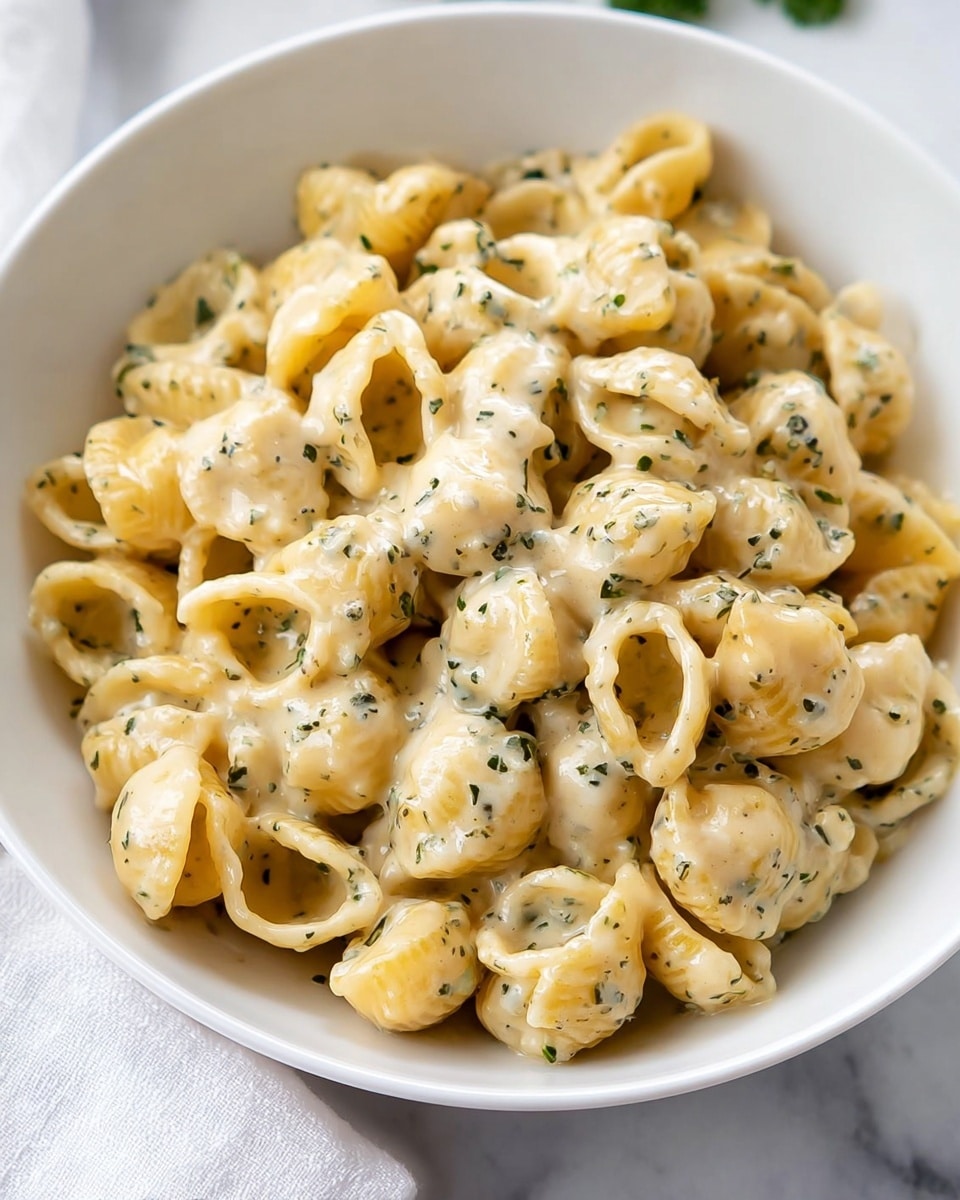 A close-up view of a white bowl filled with creamy pasta shells covered in a pale yellow sauce with herbal specks, the sauce looks smooth and coats each shell evenly. Two bright yellow lemon wedges are placed on top right inside the bowl, adding a fresh contrast. The bowl sits on a white marbled surface, and the overall colors are soft yellows and light greens from the herbs, making the dish look rich and tasty. photo taken with an iphone --ar 4:5 --v 7