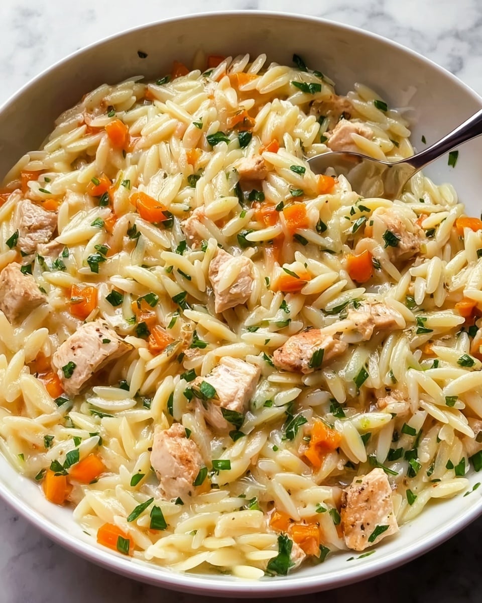 A white bowl filled with a creamy orzo pasta dish with three visible layers of ingredients mixed together: small chunks of lightly browned chicken, small diced orange carrots, and finely chopped green herbs scattered throughout. The orzo pasta pieces are plump, shiny, and white-yellowish, coated in a light sauce, creating a smooth texture that blends all the ingredients closely. A spoon is partially visible, resting inside the bowl on the upper right side. The bowl sits on a white marbled surface. photo taken with an iphone --ar 4:5 --v 7