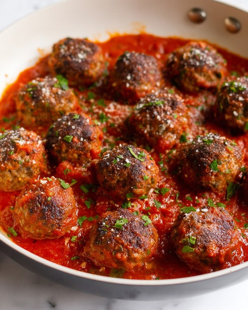 This image shows a white pan filled with about ten brownish, round meatballs that have a slightly crispy and textured surface. The meatballs sit in a thick, bright red tomato sauce that covers the bottom of the pan, with some parts showing chunks of tomato and herbs in it. The meatballs are sprinkled with small bits of white cheese and finely chopped green herbs, adding a touch of color contrast. The pan itself is close-up, filling most of the photo, and is set against a white marbled texture background. photo taken with an iphone --ar 4:5 --v 7