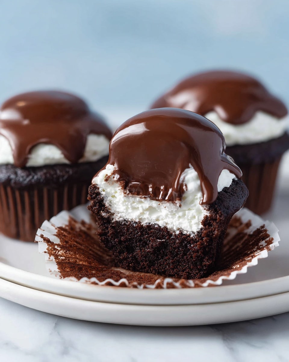 The image shows three chocolate cupcakes on a white plate placed on a white marbled surface. Each cupcake has three clear layers: a dark, moist chocolate cake base, a thick white cream layer on top of the cake, and a shiny, smooth chocolate glaze dripping slightly over the cream, forming a dome shape. One of the cupcakes in the front is partially unwrapped, exposing the crinkled white cupcake liner around the cake. The background is softly blurred with light blue tones, keeping the cupcakes as the main focus. Photo taken with an iphone --ar 4:5 --v 7