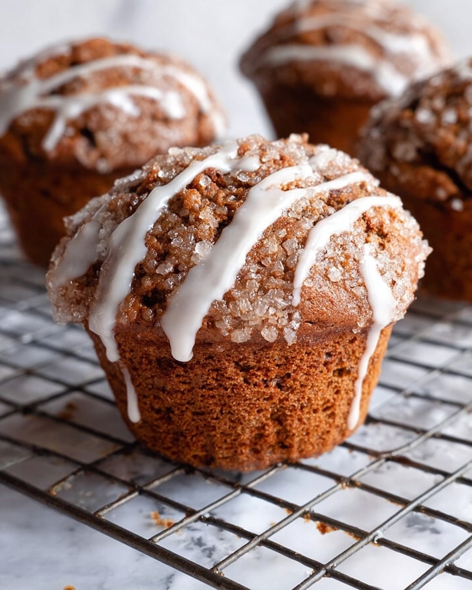 A close-up image of three brown muffins cooling on a silver wire rack above a white marbled surface, each muffin topped with a cracked textured crust covered in big sugar crystals and drizzled with white icing that partially runs down the sides; the muffins are rich and slightly rough on top with a moist look on the sides, showing a single-layer structure with the icing accentuating the textured surface. Photo taken with an iphone --ar 4:5 --v 7