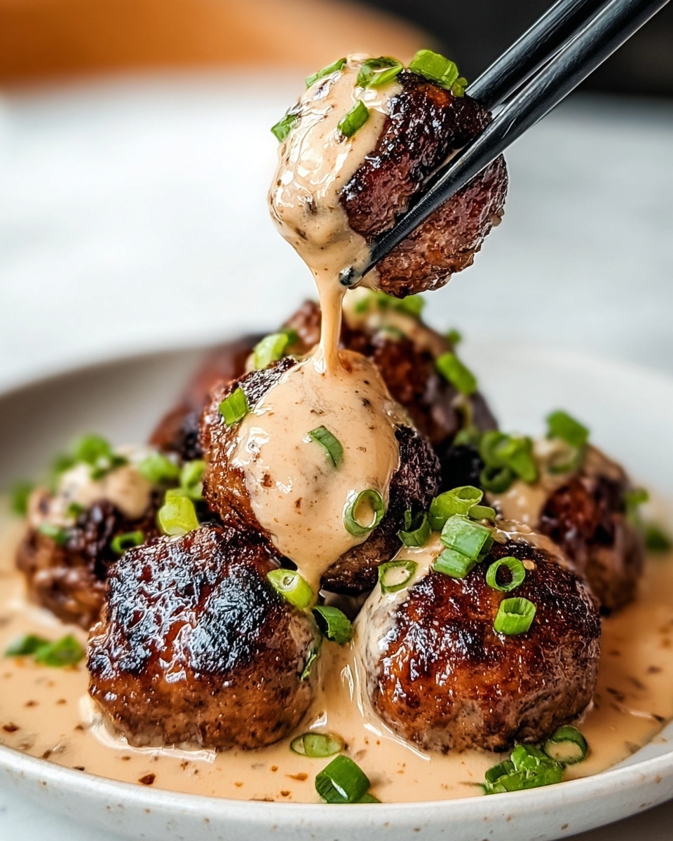 The image shows several dark brown grilled meatballs with a slightly crispy and caramelized outer texture, sitting on a shallow pool of light tan creamy sauce that has specks of seasoning throughout. The meatballs are topped with the same creamy sauce dripping down their sides, creating a rich and glossy appearance. Bright green chopped scallions are sprinkled over the meatballs and sauce, adding pops of color. One meatball is held above the plate with black chopsticks, with sauce slowly dripping from it back onto the plate. The plate is white and sits on a white marbled surface. photo taken with an iphone --ar 4:5 --v 7