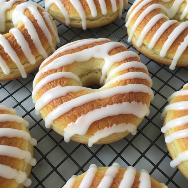 Thirteen ring-shaped donuts with a twisted swirl pattern are arranged in three rows on a black cooling rack. Each donut has a smooth, shiny glaze that gives them a light golden color with some areas slightly darker, showing the texture beneath the glossy coating. The cooling rack sits on a white marbled surface with a glimpse of a blue and white striped cloth on the left side. The donuts look soft and fresh with a consistent spiral design all around. photo taken with an iphone --ar 4:5 --v 7