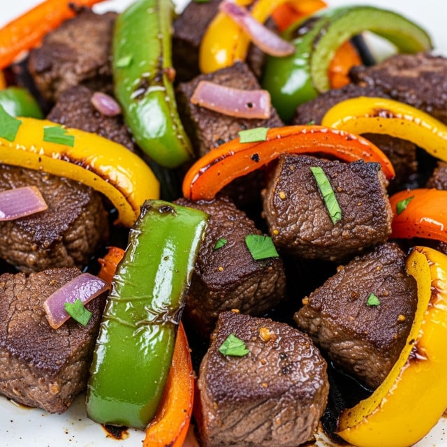The image shows a close-up view of a dish with several pieces of cooked beef and colorful bell peppers cooked together. The beef chunks appear brown and crispy on the outside, mixed with thick pieces of green, yellow, and orange bell peppers that look soft and juicy. There are also some small bits of red onion and green herbs scattered on top for color. The food is on a white surface with a slight grill texture visible underneath. The shiny, cooked food looks fresh and flavorful, with a mix of rich brown and bright bell pepper colors photo taken with an iphone --ar 4:5 --v 7