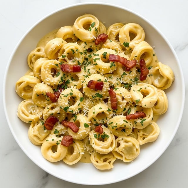 A white bowl filled with creamy tortellini pasta covered in a smooth, light yellow sauce. Small pieces of crispy, golden-brown bacon are scattered on top along with a sprinkling of finely chopped green herbs and grated cheese. The pasta rings are thick and plump, sitting closely together with visible glossy sauce coating each piece. The dish is placed on a white marbled surface. Photo taken with an iphone --ar 4:5 --v 7