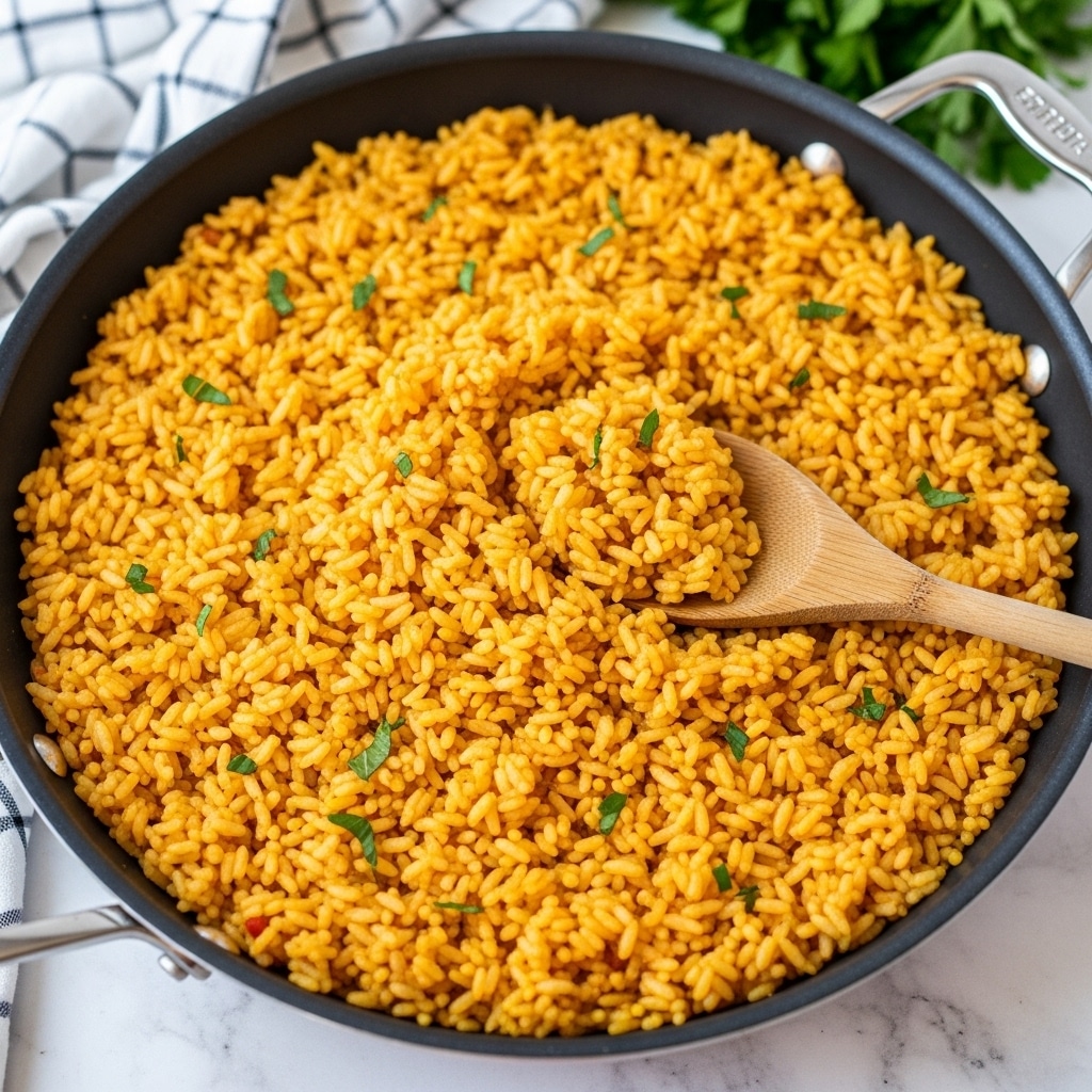 A large pan filled with a single layer of cooked yellow-orange rice, with small green herb pieces sprinkled lightly across the surface, and a wooden spoon resting in the pan, partially scooping the rice, showing its fluffy texture. The pan is placed on a white marbled surface, with a checkered cloth in the background and some green herbs slightly out of focus at the top right. The rice grains are separate and slightly glossy, giving a warm and inviting look. Photo taken with an iphone --ar 4:5 --v 7