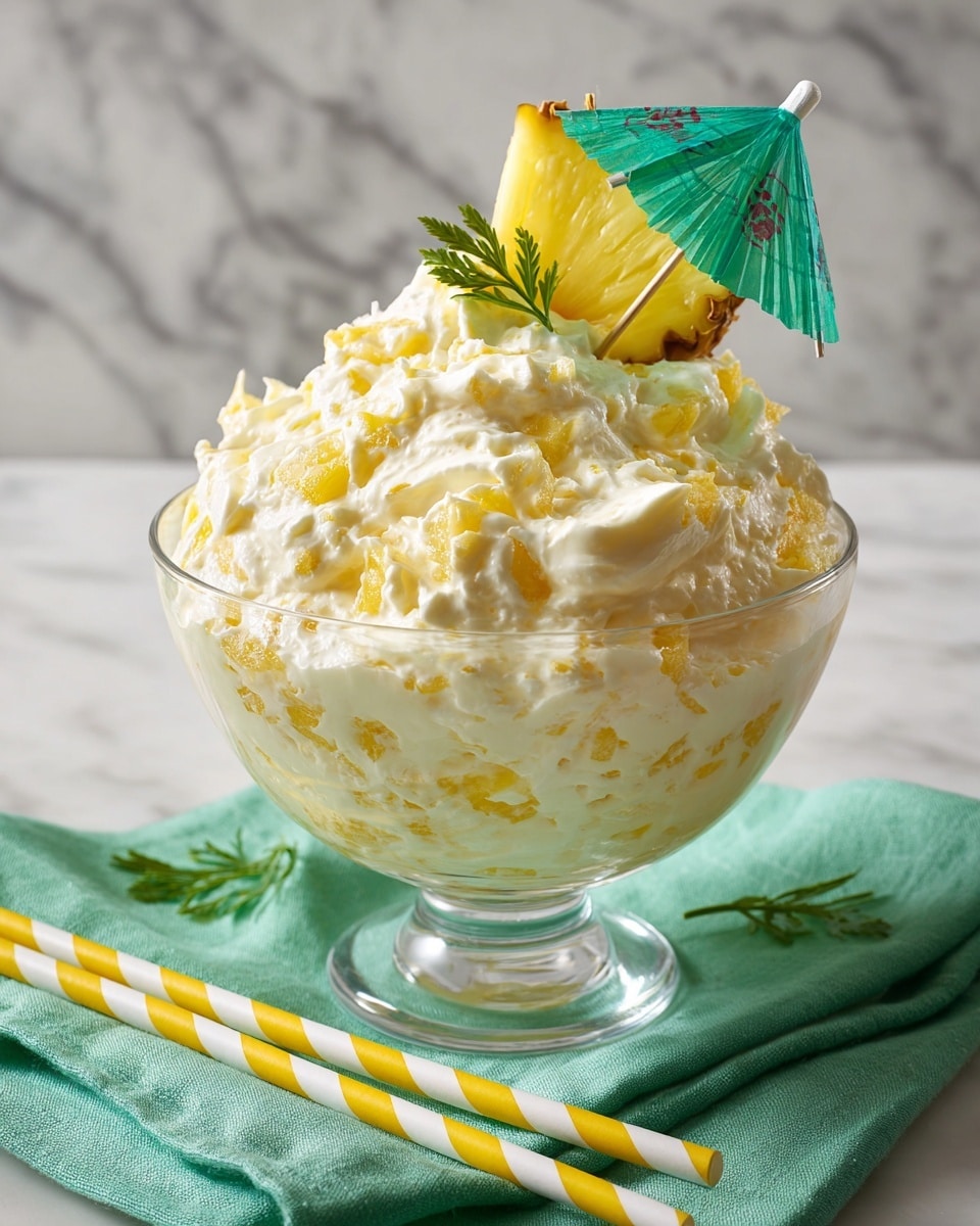 The image shows a clear glass bowl filled with a fluffy yellow and white mixture that has a creamy, whipped texture with scattered pieces of pineapple and coconut flakes mixed throughout. On top, there is a single yellow pineapple slice standing upright, adorned with a small green cocktail umbrella. The bowl is placed on a bright green cloth on a white marbled surface, with two striped straws in yellow and white resting diagonally in front. Photo taken with an iphone --ar 4:5 --v 7