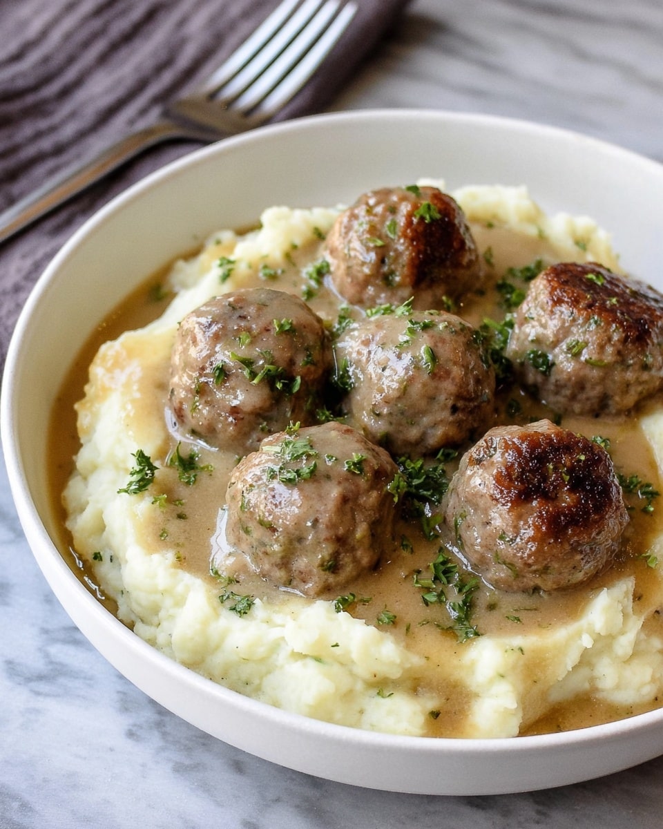 A white bowl filled with a base layer of smooth, creamy mashed potatoes in an off-white color. On top of the mashed potatoes, there are seven round meatballs, browned on the outside with a slightly textured surface. Light brown gravy covers the base around the meatballs, adding a shiny, moist look. Small green herb pieces are scattered over the meatballs and gravy, adding a touch of color contrast. The bowl sits on a white marbled texture surface with a fork blurred in the background. photo taken with an iphone --ar 4:5 --v 7