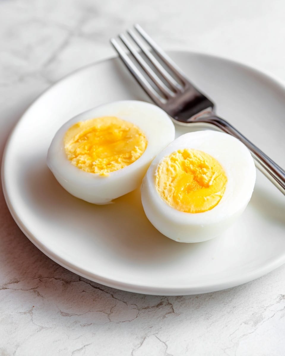 A white plate with a soft-boiled egg cut in half placed near the center. The egg's outer layer is smooth and white, while the inside shows a bright yellow yolk that is slightly runny and creamy. The two halves of the egg are touching, and the yolk texture appears soft and fresh. Behind the egg, there is a silver fork resting on the plate with its tines pointing to the upper left. The background is a white marbled texture, giving a clean and simple look to the image. Photo taken with an iphone --ar 4:5 --v 7