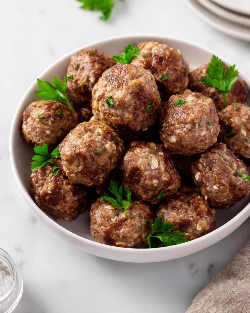 A white bowl filled with about 15 round meatballs, each with a rough texture showing bits of oats and herbs. The meatballs are brown with some green parsley leaves scattered on top and between them. The bowl sits on a white marbled surface, with another dish and a clear salt shaker partially visible on the side. The lighting is bright and clear, showing the texture of the meatballs well. photo taken with an iphone --ar 4:5 --v 7