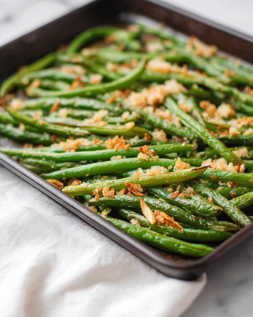 A dark baking tray filled with a single layer of green beans, each bean long and slender with a slightly wrinkled texture showing they are roasted. Scattered over the green beans are golden and light beige crispy bits of toasted garlic or bread crumbs, adding a crunchy texture on top. The tray sits on a white marbled surface with a soft white cloth partially visible beneath it. The focus is close, showing the details of the roasted green beans with some pieces browned at the edges, making the dish look fresh and tasty. photo taken with an iphone --ar 4:5 --v 7