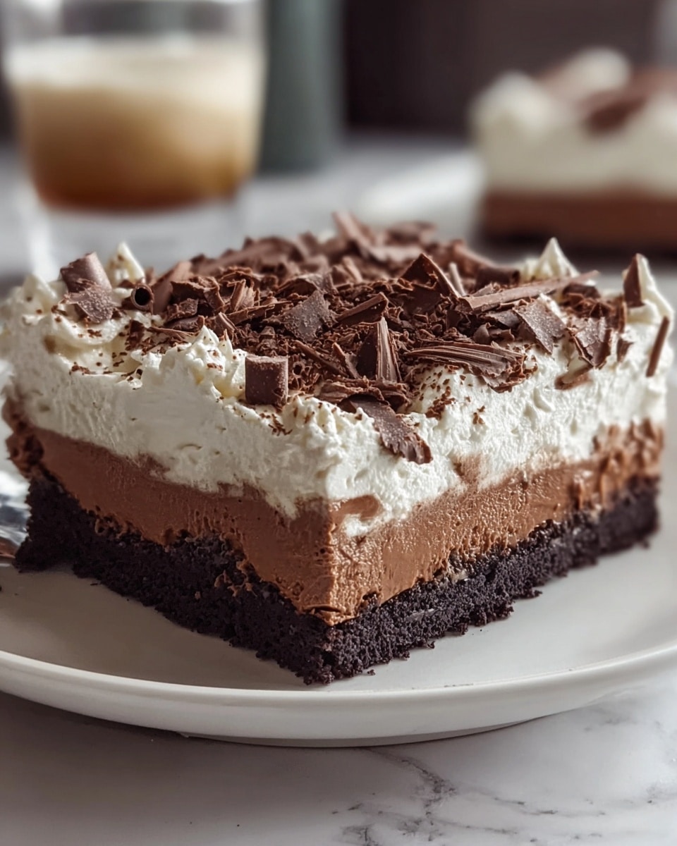 The image shows a square layered dessert on a white plate, placed on a white marbled surface. The bottom layer is a dark, crumbly chocolate crust, thick and solid. Above it is a smooth, medium brown chocolate pudding layer, thick and creamy. Next is a white whipped cream layer, fluffy and slightly thick with soft swirls around the edges. The top is covered with many irregular dark chocolate shavings scattered evenly, adding texture and a rich look. A blurred drink glass is in the background. photo taken with an iphone --ar 4:5 --v 7