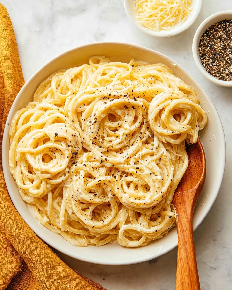 A close-up of a white bowl filled with thick, light yellow noodles covered in a creamy sauce, sprinkled with small black pepper bits and grated cheese. The noodles are loosely piled in the bowl with a wooden spoon resting on the right side, partially buried under the noodles. In the background, there are two small white dishes, one holding shredded cheese and the other cracked black pepper, all placed on a white marbled surface with a mustard yellow cloth on the bottom left corner. photo taken with an iphone --ar 4:5 --v 7