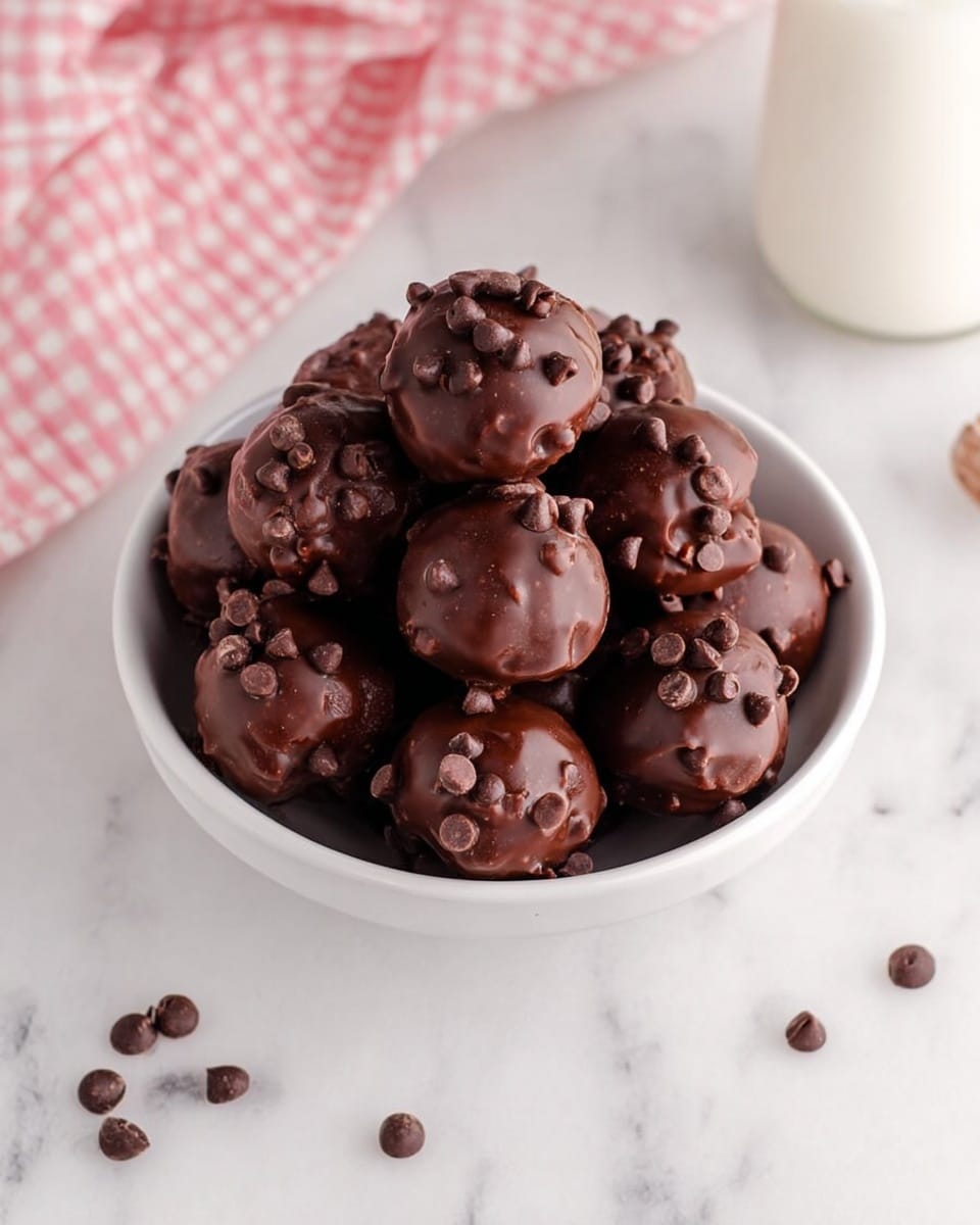 A white bowl filled with round chocolate balls, each covered in smooth dark chocolate and sprinkled with small chocolate chips on top. The chocolate balls are stacked closely, showing their shiny and slightly bumpy texture. Around the bowl, a few chocolate chips are scattered on a white marbled surface. A pink and white checkered cloth is partially visible in the top left corner, and a glass bottle of milk is on the right side in the background. photo taken with an iphone --ar 4:5 --v 7