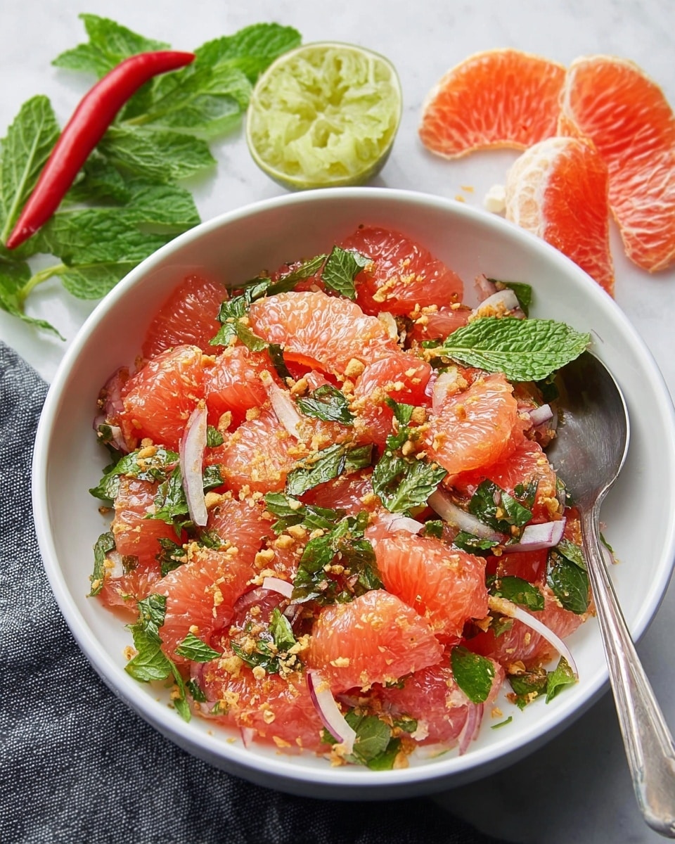 A white bowl filled with a colorful salad made of bright pink grapefruit chunks and thinly sliced pale purple onions mixed with fresh green mint and cilantro leaves, topped with small crushed nuts or crunchy bits. Nearby are half a cut lime, a red chili pepper, a few mint leaves, and peeled grapefruit segments on a white marbled surface. A shiny silver spoon rests inside the bowl. Photo taken with an iphone --ar 4:5 --v 7