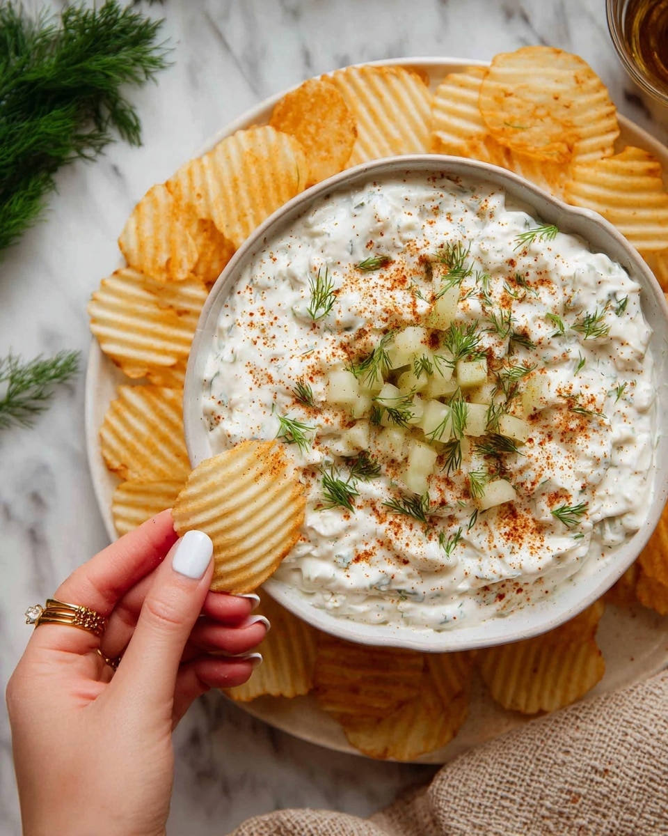A white bowl filled with a creamy white dip topped with small green dill leaves, tiny pale green pickle pieces, and a dusting of reddish-brown seasoning evenly spread across the top in a circular pattern. The bowl is placed on a white plate surrounded by ridged potato chips. A woman's hand with a gold ring and neatly shaped white nails is holding a ridged potato chip dipped into the creamy dip. The surface beneath is a white marbled texture, with some green dill branches and a beige textured cloth visible on the side. photo taken with an iphone --ar 4:5 --v 7