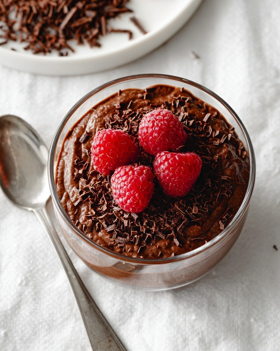 A clear glass bowl filled with a rich, dark brown chocolate pudding with a slightly bumpy texture, topped with small, dark chocolate shavings spread in the middle and four bright red raspberries with frosty details arranged closely on top. The bowl is placed on a white marbled textured cloth. A shiny silver spoon is positioned to the left side of the bowl. At the top, part of a white plate with more dark chocolate shavings is partially visible. photo taken with an iphone --ar 4:5 --v 7