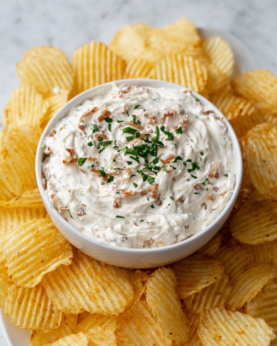 A white bowl filled with creamy onion dip that has a thick, smooth texture with small bits of browned onion visible throughout, garnished with chopped fresh green herbs on top. The bowl sits in the center of a pile of golden yellow ridged potato chips arranged around it. The whole scene is on a white marbled surface. photo taken with an iphone --ar 4:5 --v 7