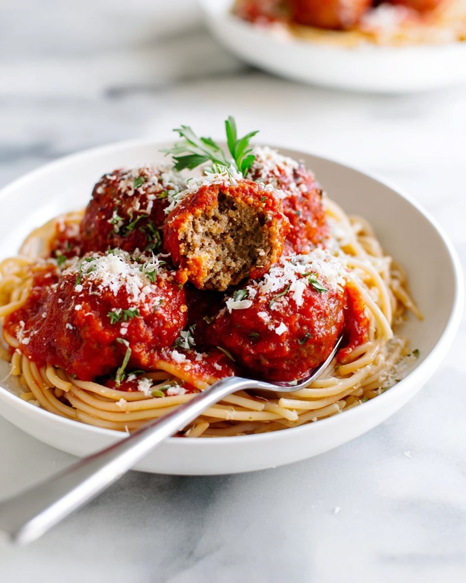 A white plate holds a neat nest of light brown spaghetti at the bottom layer, topped with several round meatballs covered in bright red tomato sauce. One meatball is cut open, showing a dense, crumbly brown inside. The red sauce is thick and chunky, spilling slightly over the pasta. White grated cheese is sprinkled generously on top of the meatballs and sauce, with a small green herb sprig placed as garnish. A silver fork rests on the plate, partially inserted into the pasta. The background has a white marbled texture, with soft lighting bringing out the colors vividly. Photo taken with an iphone --ar 4:5 --v 7