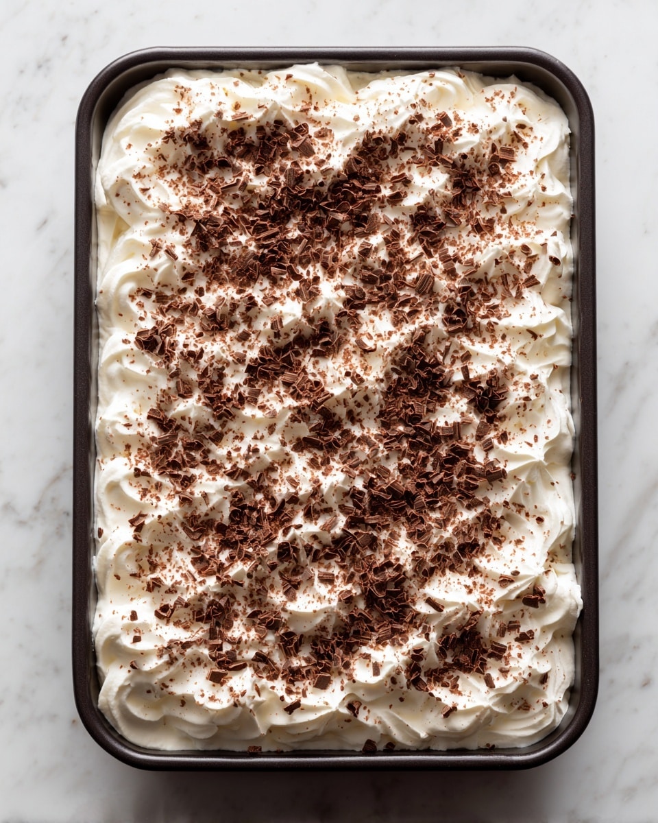 A slice of dessert on a white plate showing three clear layers: a bottom dark brown crumbly crust, a middle smooth light brown creamy layer, and a top fluffy white cream layer sprinkled with small chocolate shavings. Next to the plate, there is a silver fork resting on it. In the background, a white rectangular dish with the rest of the dessert, showing the same three layers, sits on a white marbled surface with a yellow and white striped cloth partially under the dessert dish. Photo taken with an iphone --ar 4:5 --v 7