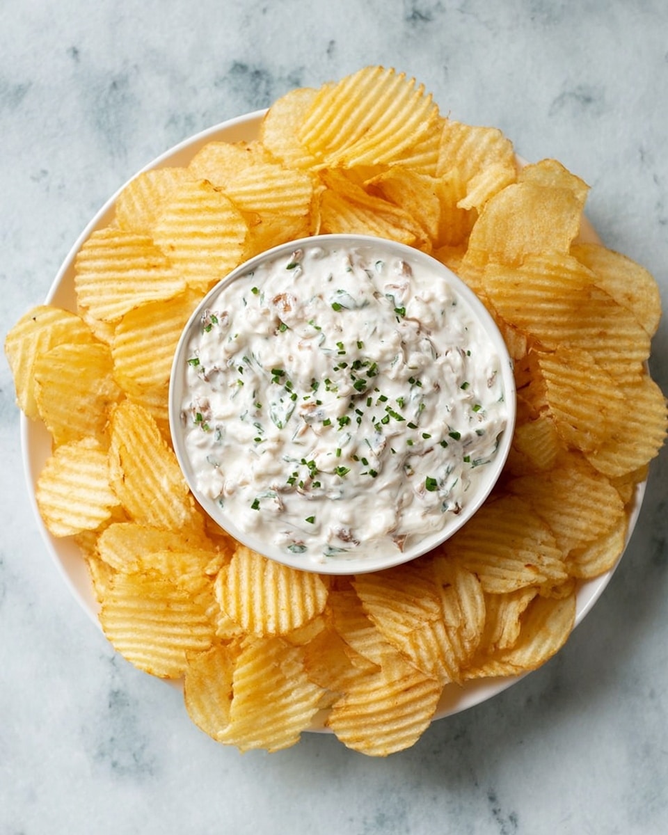 A white bowl of thick white dip with small brown bits mixed in is placed in the center of a white plate filled with golden-yellow ridged potato chips arranged around it. The dip is topped with small pieces of green herbs. All of this is set on a white marbled textured background. photo taken with an iphone --ar 4:5 --v 7
