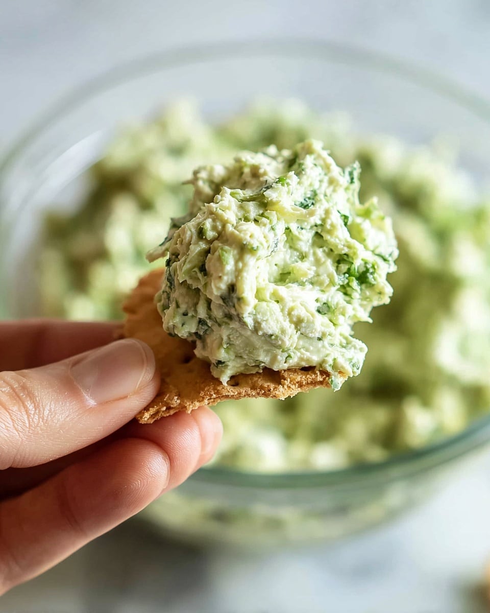 A close-up of a woman's hand holding a piece of light brown cracker topped with a thick scoop of chunky, pale green spread mixed with small bits of darker green herbs. In the background, there is a clear glass bowl filled with the same green spread, all set on a white marbled surface. The texture of the spread looks creamy yet slightly coarse with visible herb pieces. photo taken with an iphone --ar 4:5 --v 7